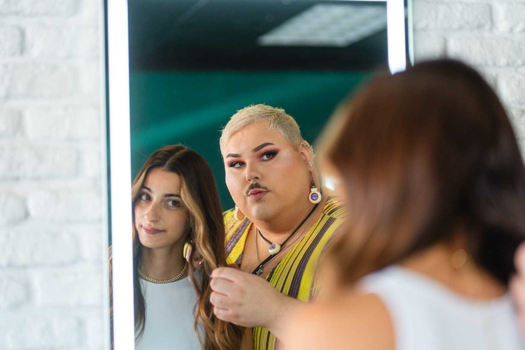 Two people styling hair in a salon, with one holding a hairbrush, reflecting in a mirror.
