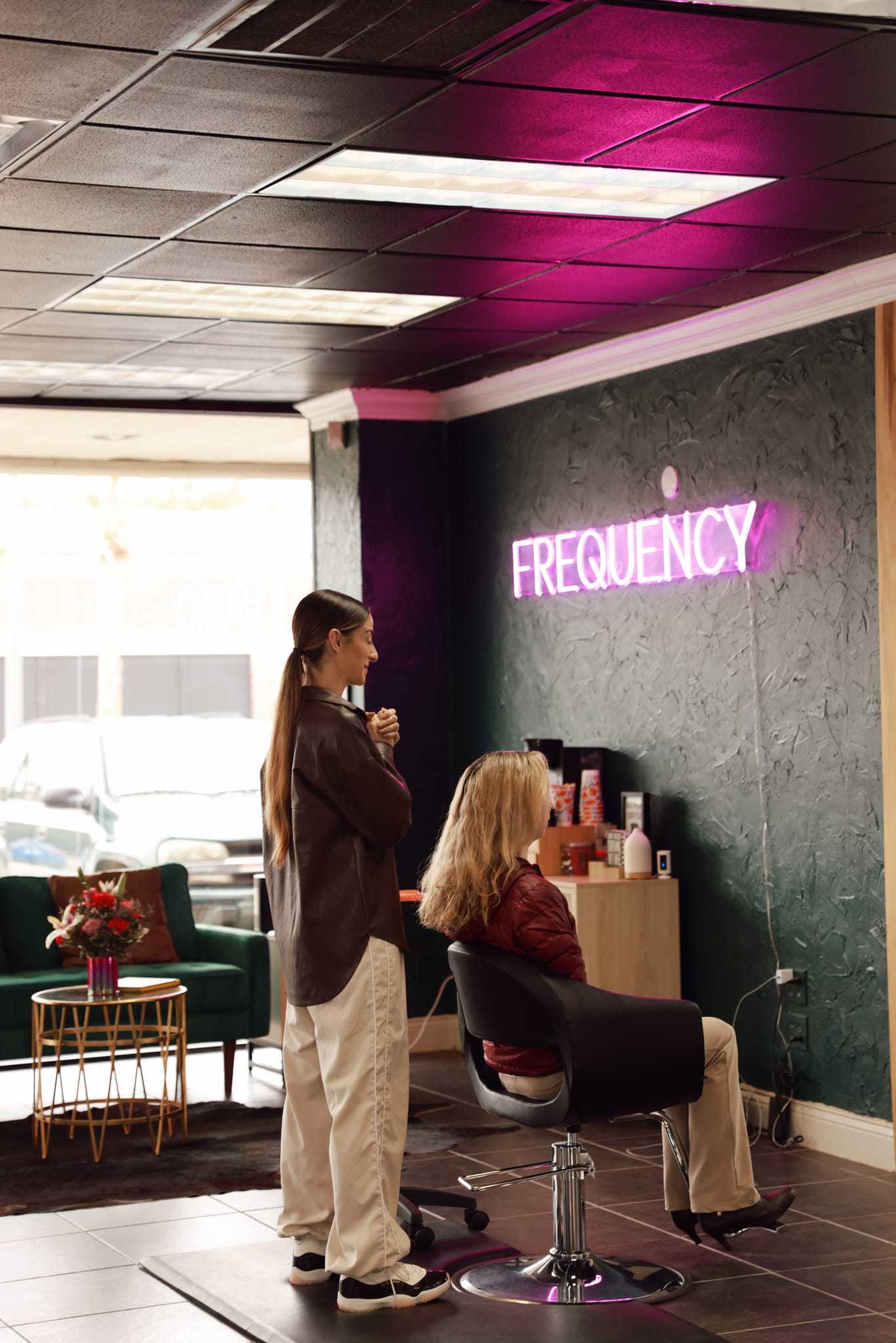 Salon interior with neon "FREQUENCY" sign and two people, one seated, under pink lighting.